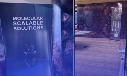 Entrada de um evento da Hologic em Porto, Portugal. Um roll up banner azul exibe os textos “BEM-VINDOS” e “MOLECULAR SCALABLE SOLUTIONS”, enquanto uma porta aberta revela um espaço decorado. Na parte inferior, lê-se “Hologic Molecular Solutions Day - Porto, Portugal - 26 de Novembro 2025”.